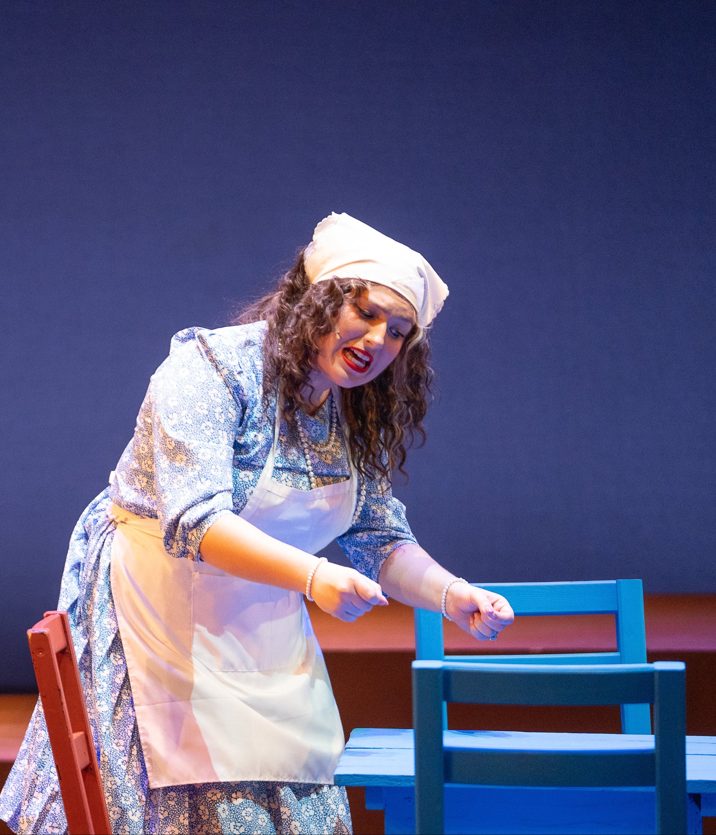 Woman in a theatrical performance with a blue chair and red bench on a dark background